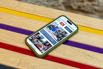 Smartphone zeigt die Website des Lokschuppens Rosenheim. Es liegt auf einer bunten Holzbank.