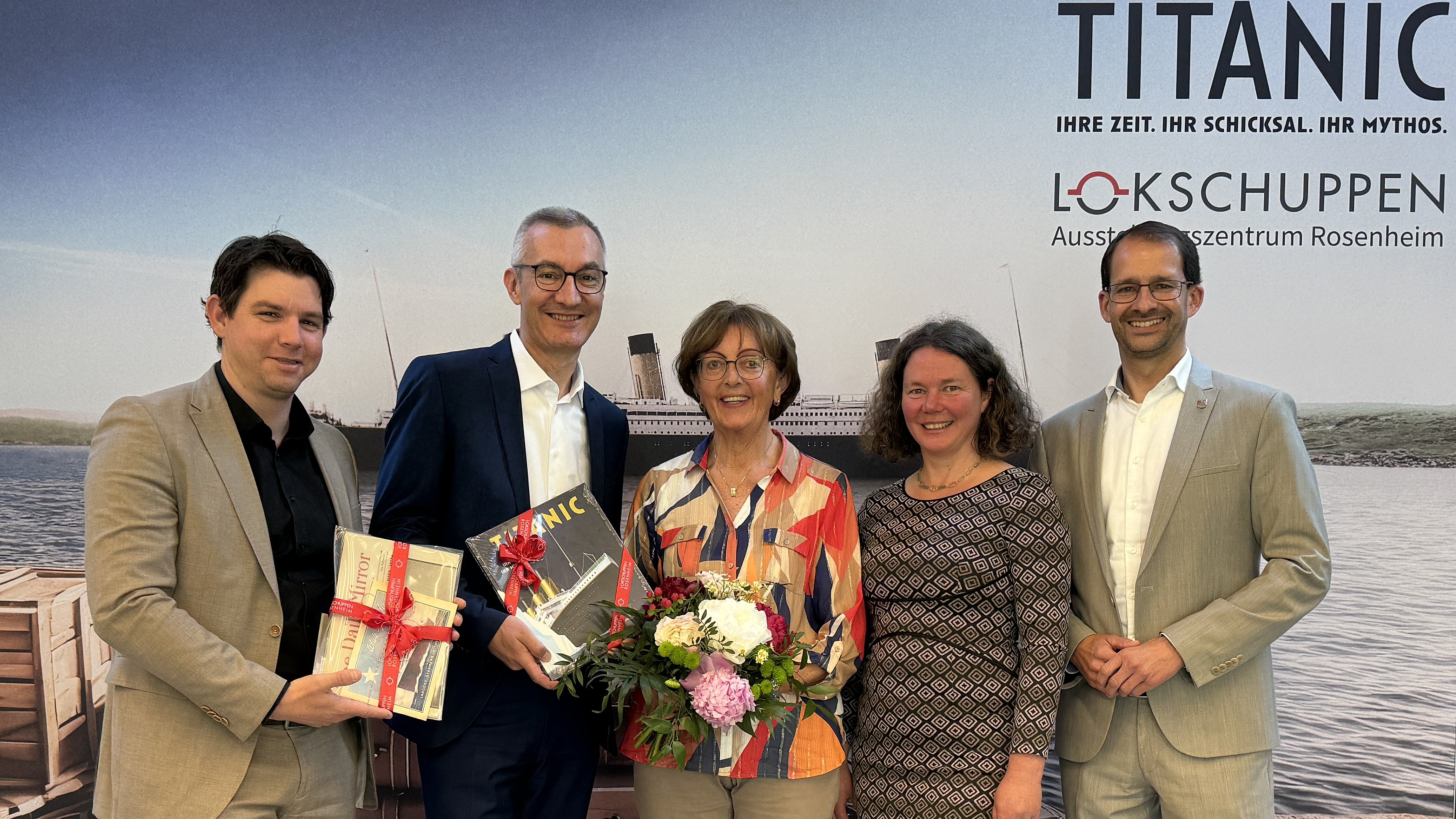 Fünf Personen stehen gemeinsam vor einem Titanic-Ausstellungsplakat und halten Blumen und Geschenke.