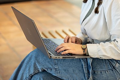 Person sitzt auf einer Holzbank und tippt auf einem Laptop in entspannter Sitzhaltung.