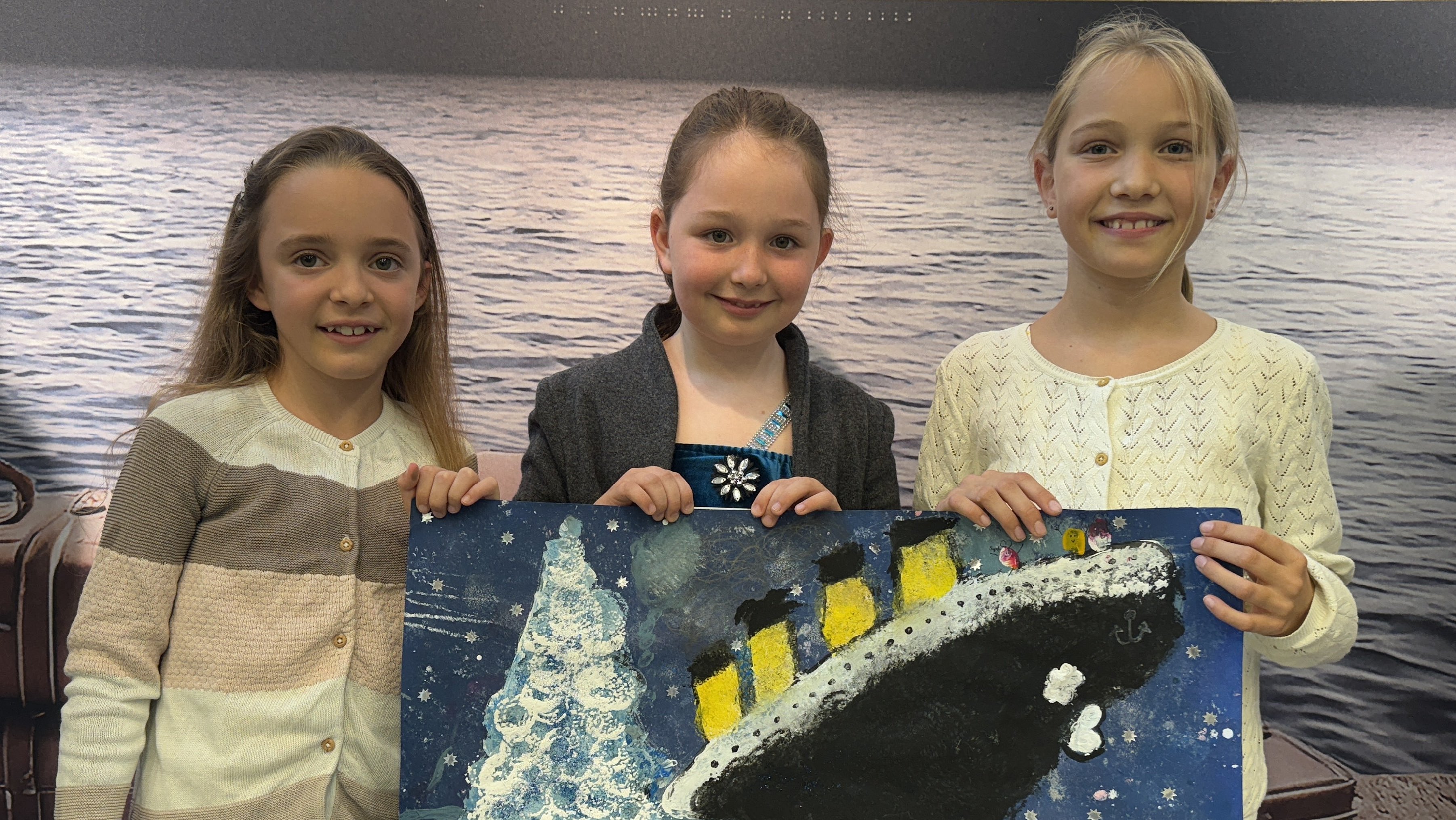 Drei Grundschülerinnen halten ihr Gewinnerbild des Titanic-Malwettbewerbs, das die sinkende Titanic und einen Eisberg zeigt.