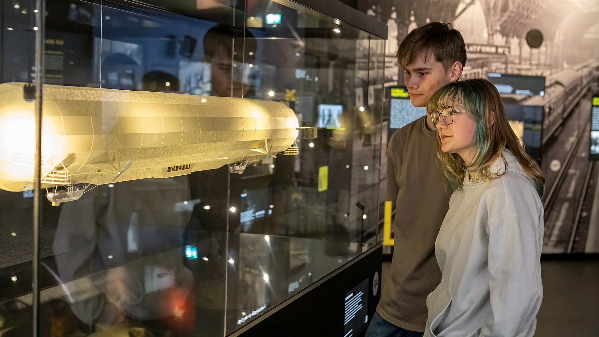 Zwei junge Erwachsene betrachten in der TITANIC-Ausstellung ein beleuchtetes Modell eines Zeppelins in einer Vitrine.