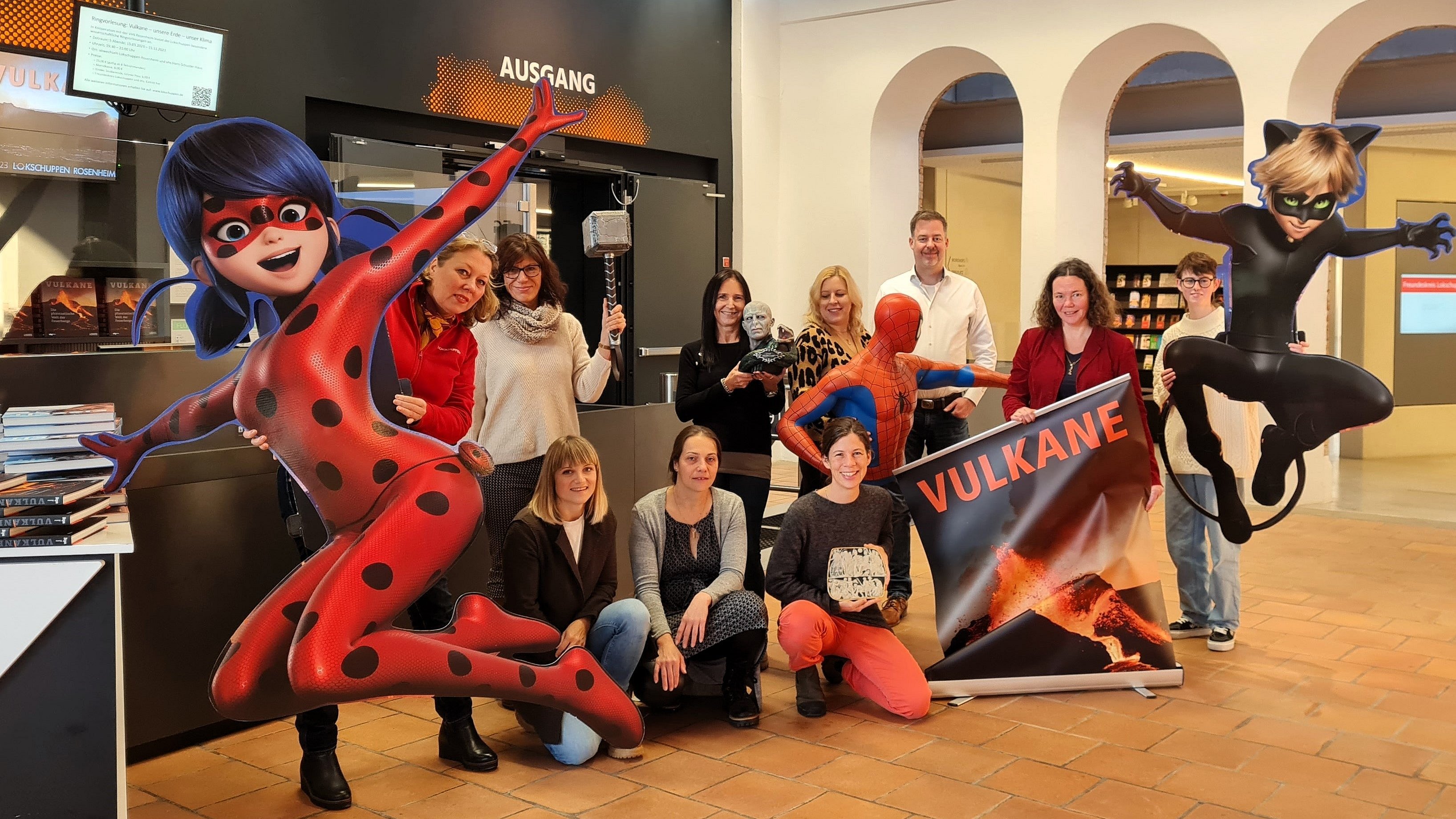 Das zehnköpfige Lokschuppen-Team mit Jennifer Morscheiser posiert im Foyer, flankiert von großen Figuren der Superhelden Ladybug, Spiderman und Cat Noir.