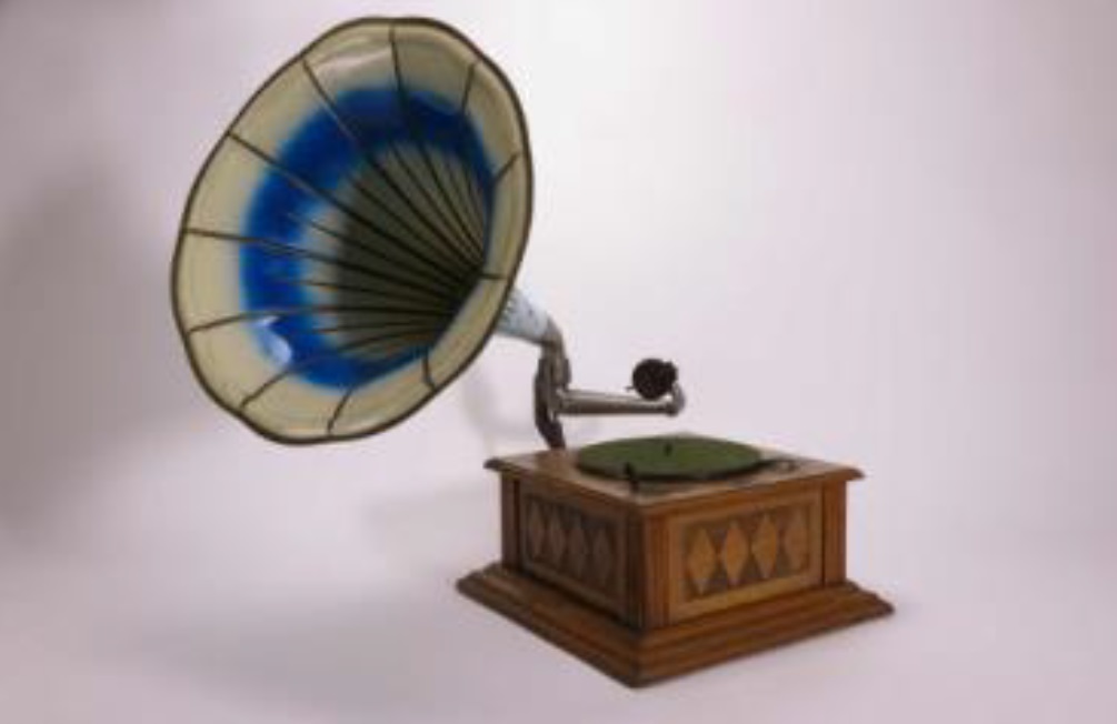 Ein historisches Grammophon mit großem, trichterförmigem Lautsprecher. Der Schalltrichter ist innen blau und außen hell, das Gehäuse ist aus Holz mit dekorativem Muster. Das Gerät steht auf einem hellen Untergrund.
