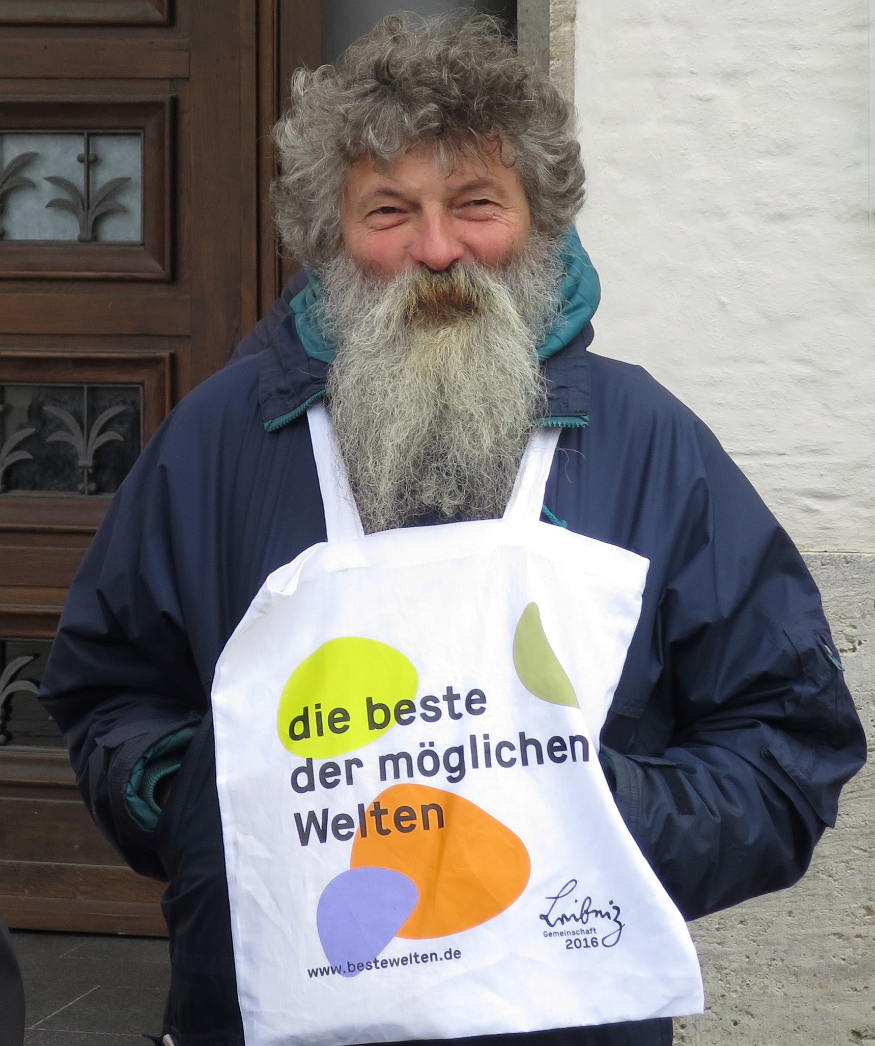 Älterer Mann mit grauem Vollbart trägt eine weiße Stofftasche mit bunter Aufschrift.