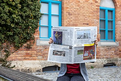 Person liest eine Zeitung vor dem Lokschuppen Rosenheim. Im Hintergrund ist das Efeu-bewachsene Backsteingebäude mit blauen Fensterrahmen zu sehen.